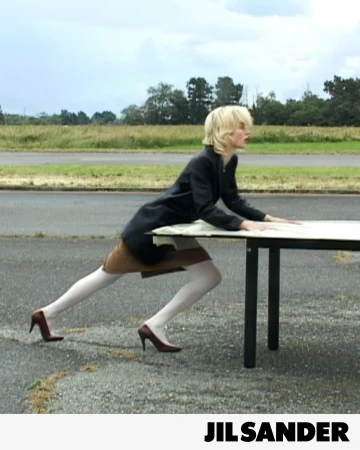 Model pushing a large table across an open paved airfield runway wearing a black coat and white tights for Jil Sander.