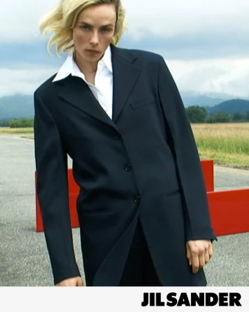 Model Edie Campbell wearing a tailored black coat and white collared shirt outdoors against a cloudy sky.