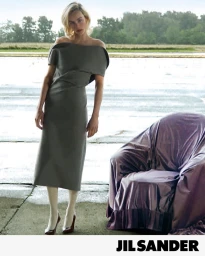 Edie Campbell in a structured grey off-the-shoulder dress standing beside a purple-draped armchair outdoors for Jil Sander.