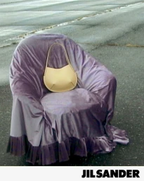Jil Sander beige structured shoulder bag placed on a purple velvet-draped armchair on an asphalt lot.