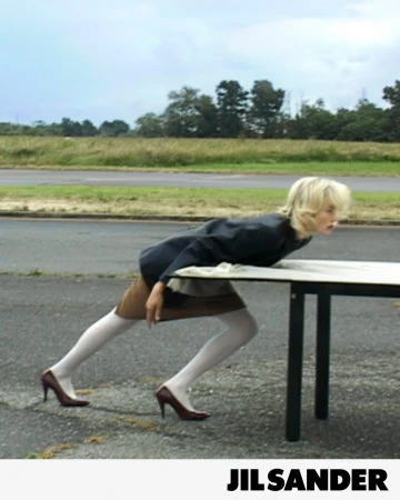 Model Edie Campbell wearing a dark jacket, beige skirt, white tights, and burgundy heels leans forward over a table on an open asphalt road.