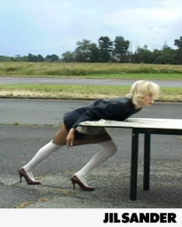 Model Edie Campbell wearing a dark jacket, beige skirt, white tights, and burgundy heels leans forward over a table on an open asphalt road.