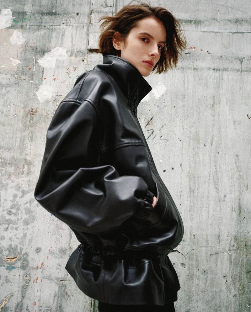 Model with short brown hair looking over her shoulder wearing an oversized black leather jacket against a weathered concrete wall.