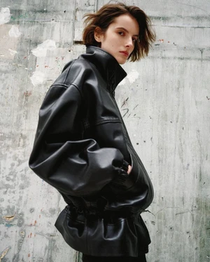 Model wearing an oversized high-collar black leather jacket posed against a textured grey concrete wall.