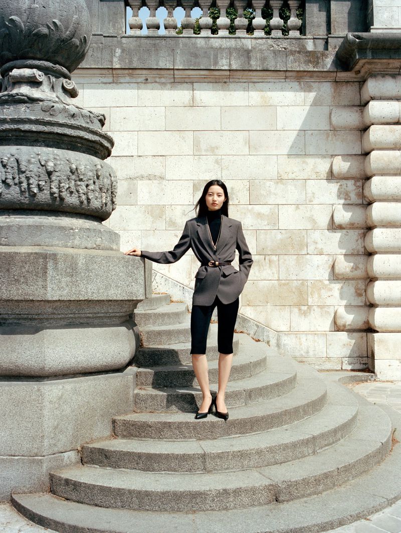 Model Chen Yang posing in a grey belted blazer and black knee-length shorts on classical stone stairs for Aritzia.