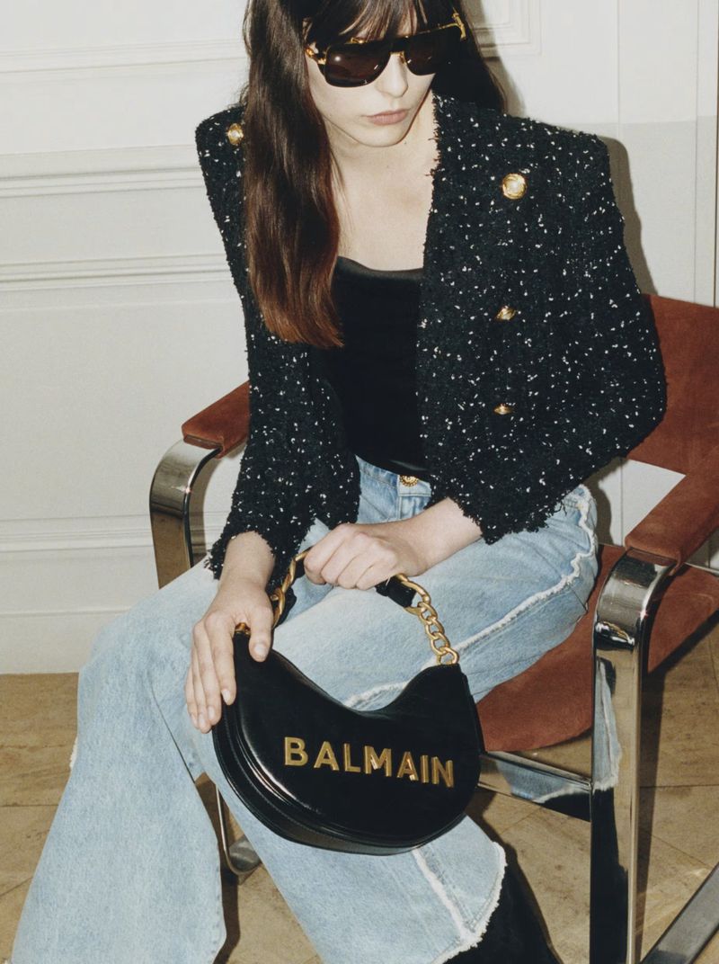 Woman in tweed blazer and light jeans sits holding a black Balmain handbag in an indoor setting, showcasing fashion and style.