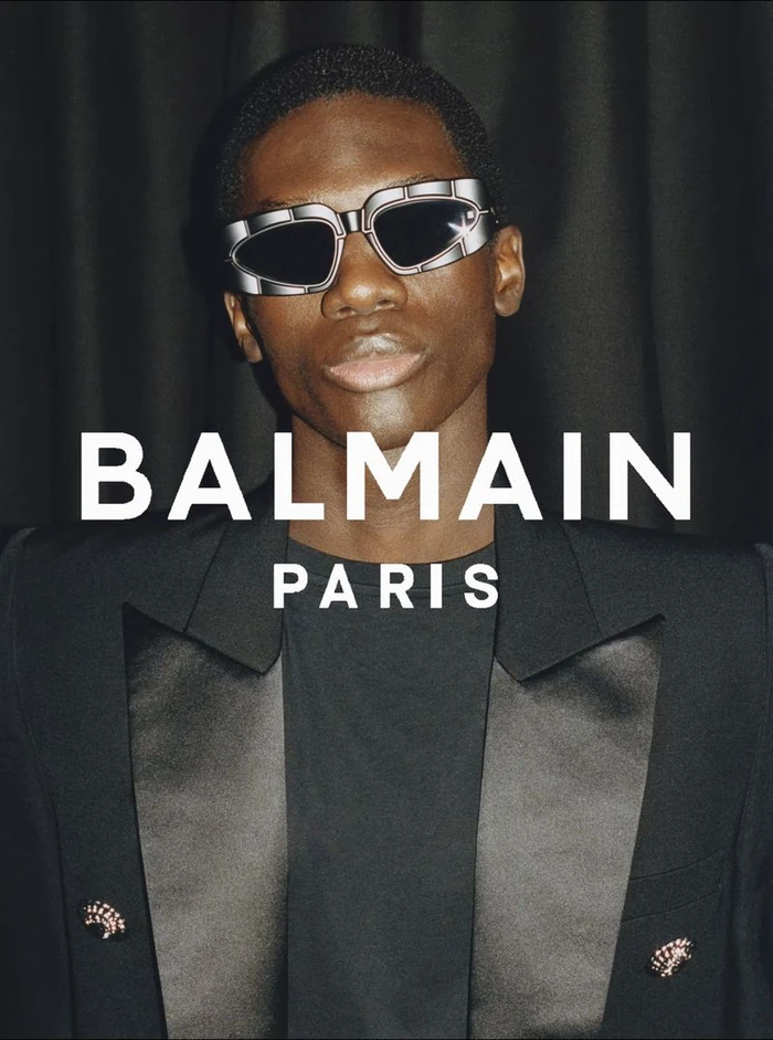 Model wearing silver futuristic sunglasses and a black blazer with satin lapels against dark fabric