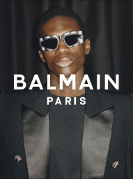 Model wearing silver futuristic sunglasses and a black blazer with satin lapels against dark fabric