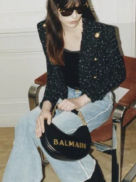 Model seated wearing a black tweed jacket, light wash jeans, sunglasses, and holding a black handbag with gold logo lettering