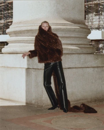 Maggie Maurer wears a brown textured faux-fur jacket and black leather trousers, posing against a massive stone pillar.