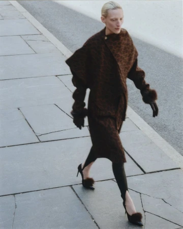 Model Maggie Maurer walks on a paved sidewalk wearing a textured brown coat with fur cuffs, stirrup leggings, and fur-accented heels.