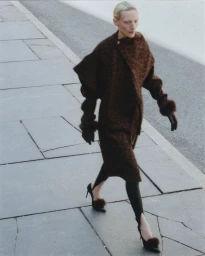 Model Maggie Maurer walks on a paved sidewalk wearing a textured brown coat with fur cuffs, stirrup leggings, and fur-accented heels.