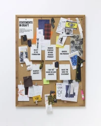 A framed cork pinboard with typographic cards, fabric swatches, sketches, and an AirWair tag mounted on a white background.
