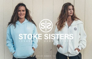 Two women wearing light blue and off-white hoodies standing against a vertical wooden wall for USBR Apparel Stoke Sisters.
