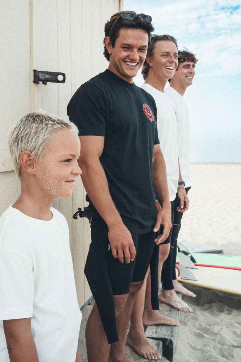 Group of four smiling surfers of various ages lined up outside a wooden beach shack on the sand with surfboards.