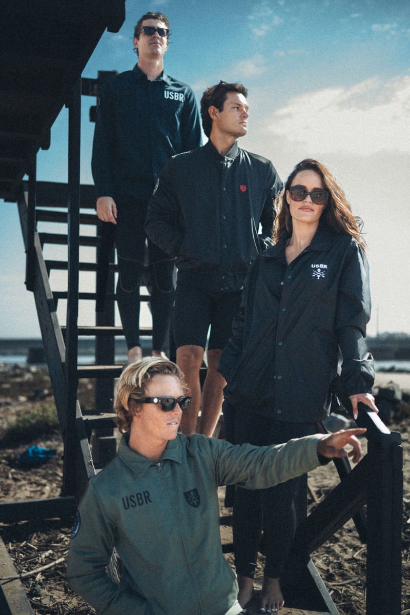 Four models posing on a black wooden staircase near a beach wearing USBR Apparel jackets and sunglasses.