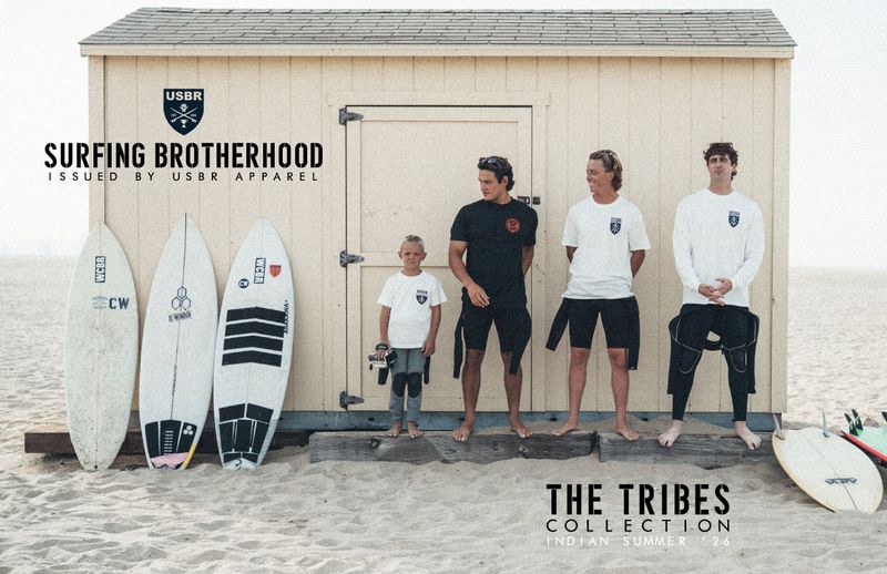Four surfers of different ages standing with surfboards outside a wooden beach shack on sand for USBR Apparel.
