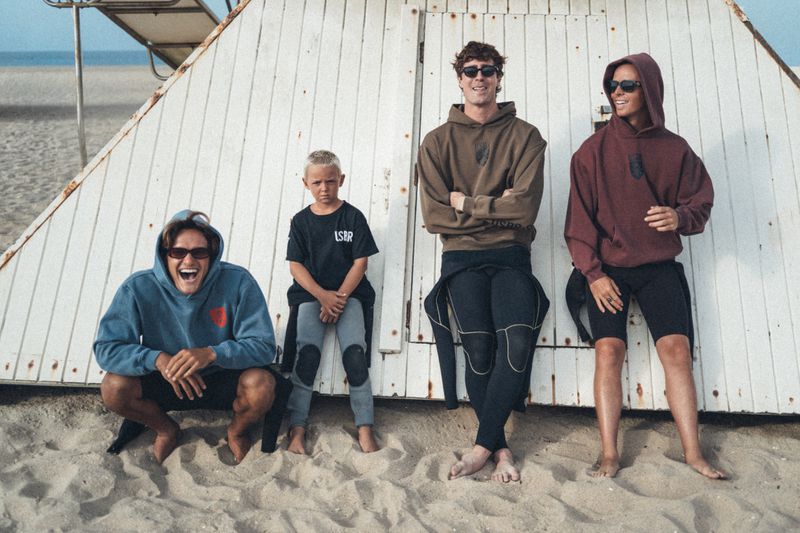 Four males of varying ages wearing casual hoodies and wetsuits posing in front of a white wooden beach hut on the sand.