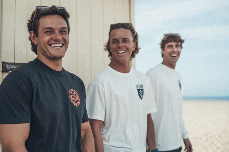 Three smiling young men wearing USBR graphic t-shirts outdoors next to a beach hut with sand in the background.