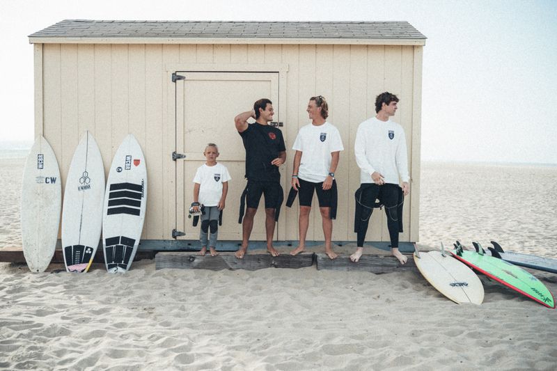 Four male surfers of varying ages standing outside a cream wooden beach hut on a sandy beach surrounded by surfboards.