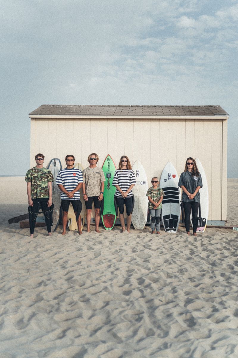 Group of six surfers standing on a sandy beach with surfboards against a wooden beach hut under a pale blue sky.
