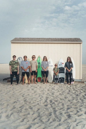Group of six surfers standing on a sandy beach with surfboards against a wooden beach hut under a pale blue sky.