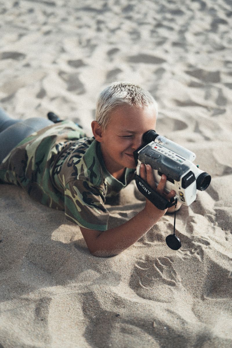 Young boy with blonde hair lying on a sandy beach holding a vintage camcorder to his eye in a camouflage t-shirt.