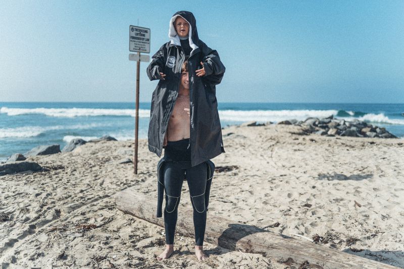 Two people stacked in a playful pose wearing an oversized black hooded surf robe on a sandy beach by the ocean.