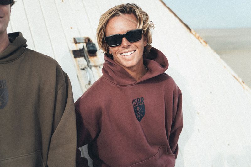 A smiling young blonde man wearing black sunglasses and a burgundy USBR hooded sweatshirt outdoors beside a companion.