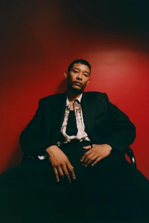Model seated in a black tailored suit jacket and patterned shirt against a vibrant red backdrop for Public School.
