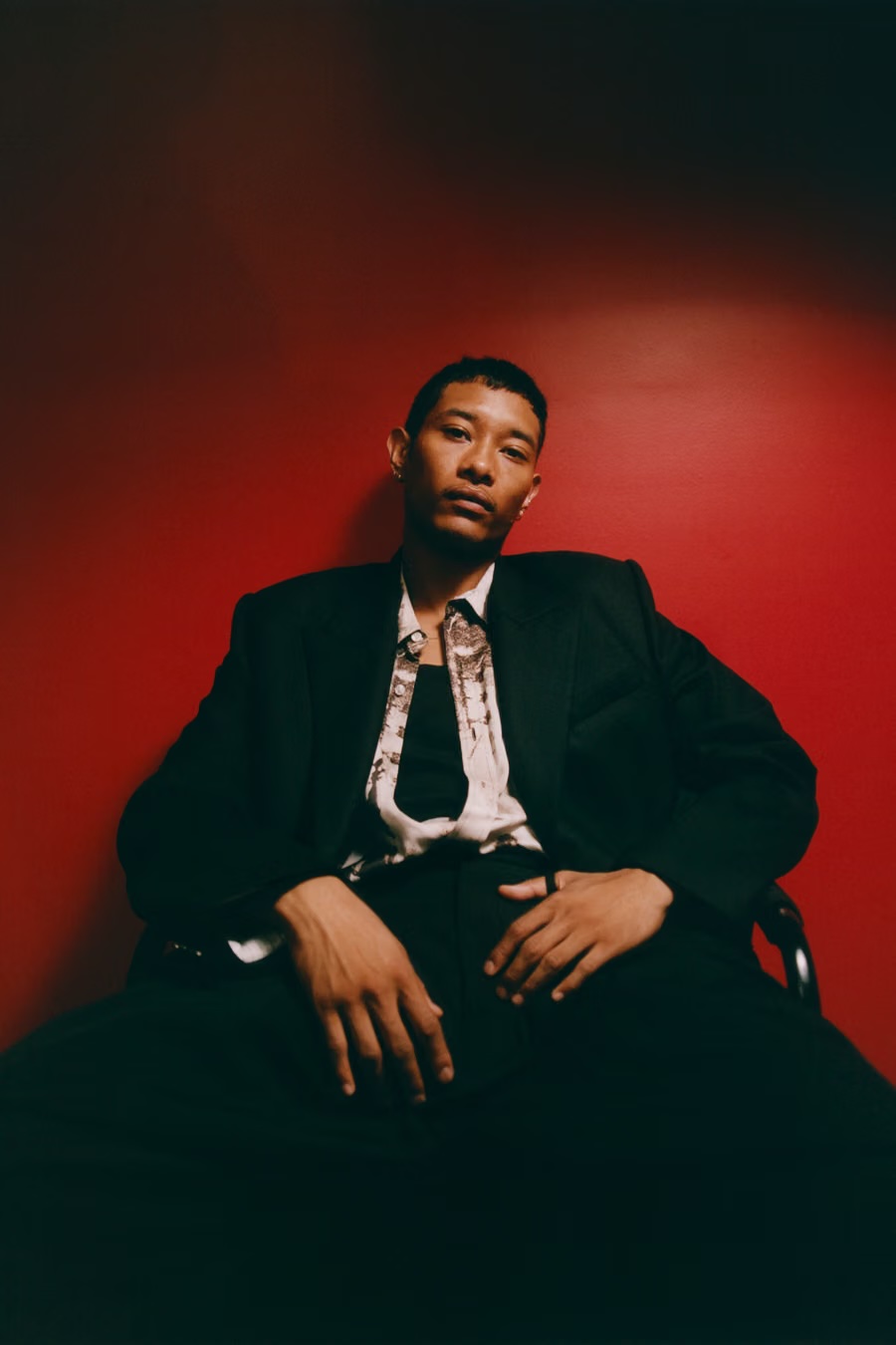 Seated man wearing a black blazer and patterned open shirt against a deep red backdrop in low light.