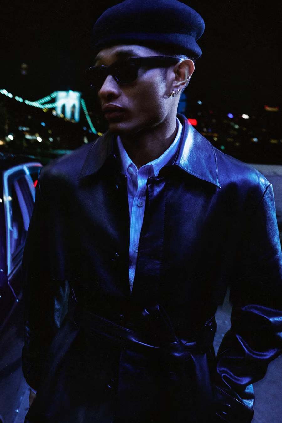 Model wearing dark sunglasses, a black beret, and a black leather coat standing outdoors at night with an illuminated bridge behind.