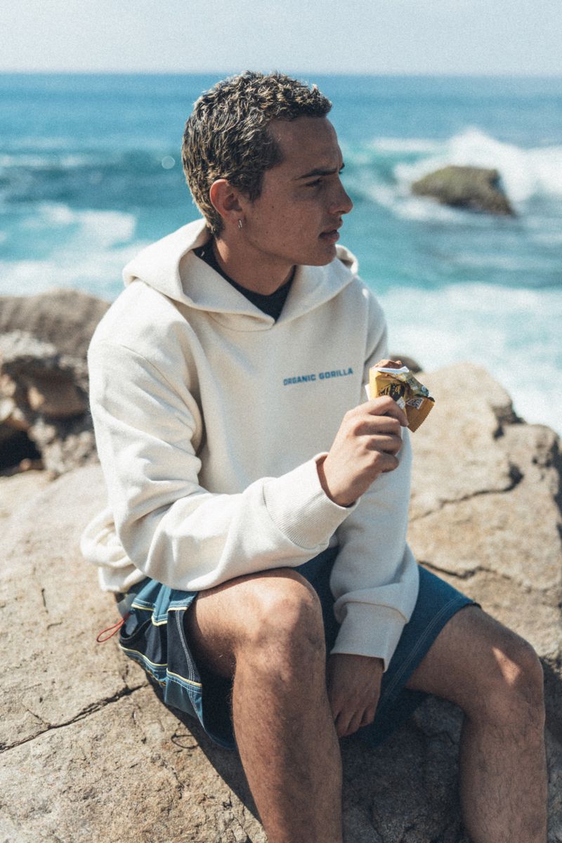 A young man sitting on rocky coastal terrain wearing a cream hoodie and blue shorts while holding a wrapped snack.