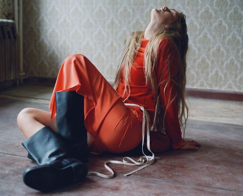 Model Iris Lasnet in a bright orange long-sleeve dress and black leather boots seated on a wooden floor against patterned wallpaper.
