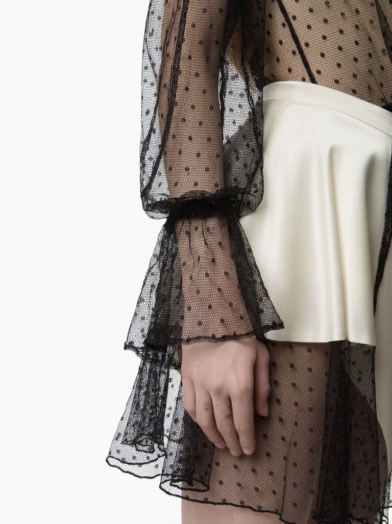 Close-up of a person wearing a black polka dot mesh long sleeve top and a white skirt, showcasing fashion and design details.