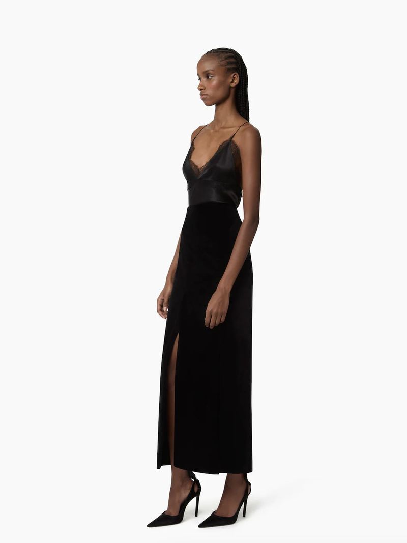 Woman poses in black silk camisole, velvet slit skirt, and stiletto heels against white background in side view in a fashion shot.