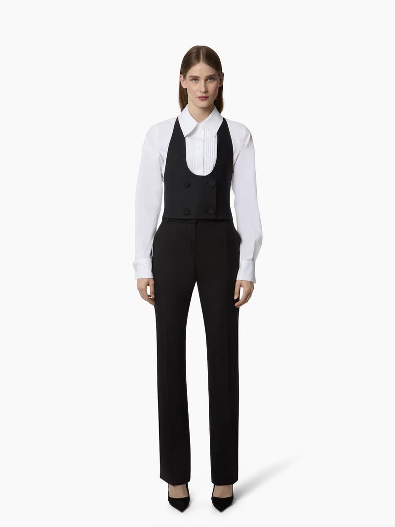 Fashion shot of a woman wearing a white shirt, black vest, black pants, and black high heels, posing in front of a white background.