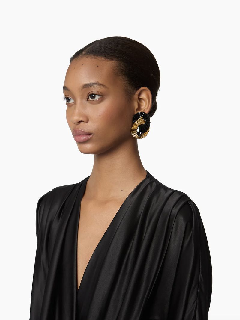 Portrait of an adult female wearing statement earrings and a black dress against a white backdrop in a studio setting.