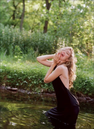 Gigi Hadid wading in a forest stream wearing a black backless dress with her hands raised to her face.