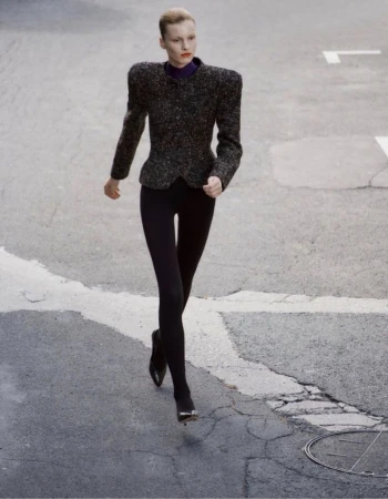 Model walking forward on an asphalt street in an exaggerated shoulder tweed jacket and black leggings.