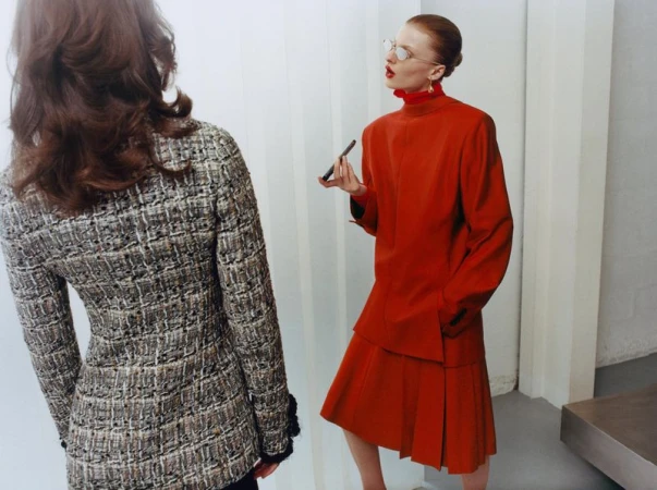 Model in a structured red coat and pleated skirt holds a phone while standing opposite a woman in a grey tweed jacket.