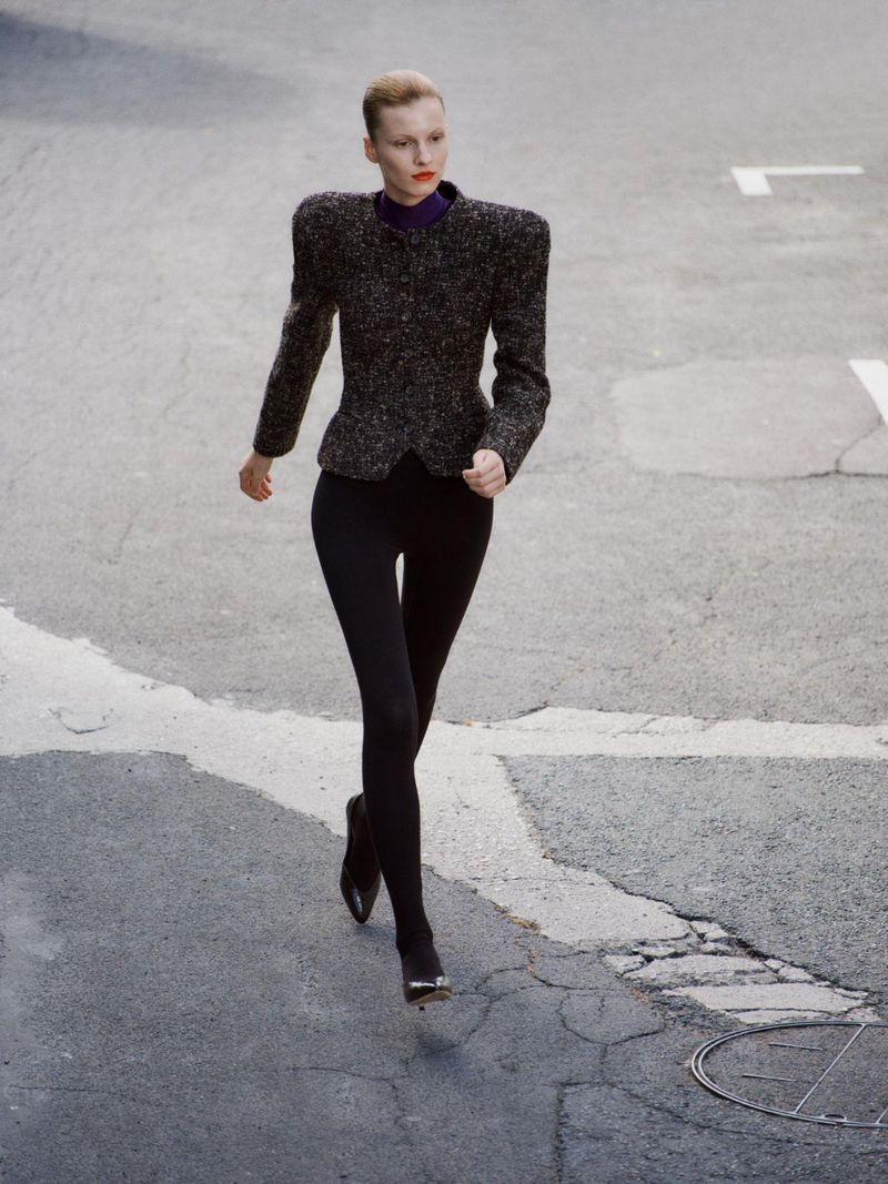 Model walking outdoors in a broad-shouldered textured grey jacket, black leggings, and heels on an asphalt street.