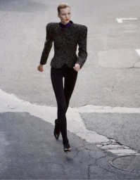 Fashion model walking on an asphalt street wearing an exaggerated shoulder tweed jacket, black leggings, and pointed high heels.