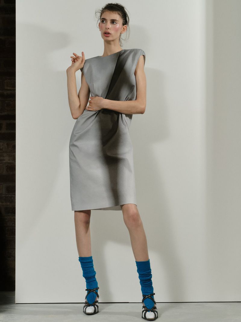 A fashion model in a structured grey shift dress, bright blue socks, and strappy sandals posing against a white studio wall.