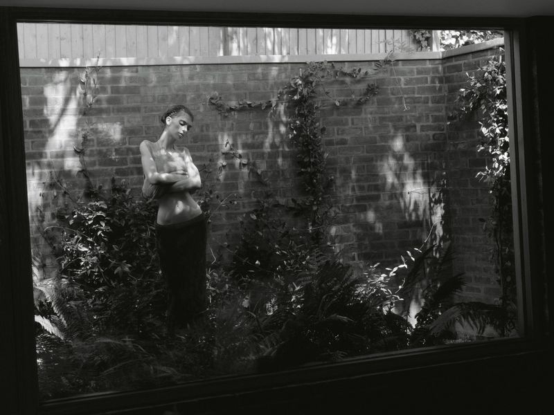 Monochrome shot of a model standing among ferns in front of a brick wall, framed through a wide window.
