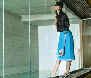 Fashion model in a black satin top and bright blue skirt standing beside glass panels over a modern indoor pool area.