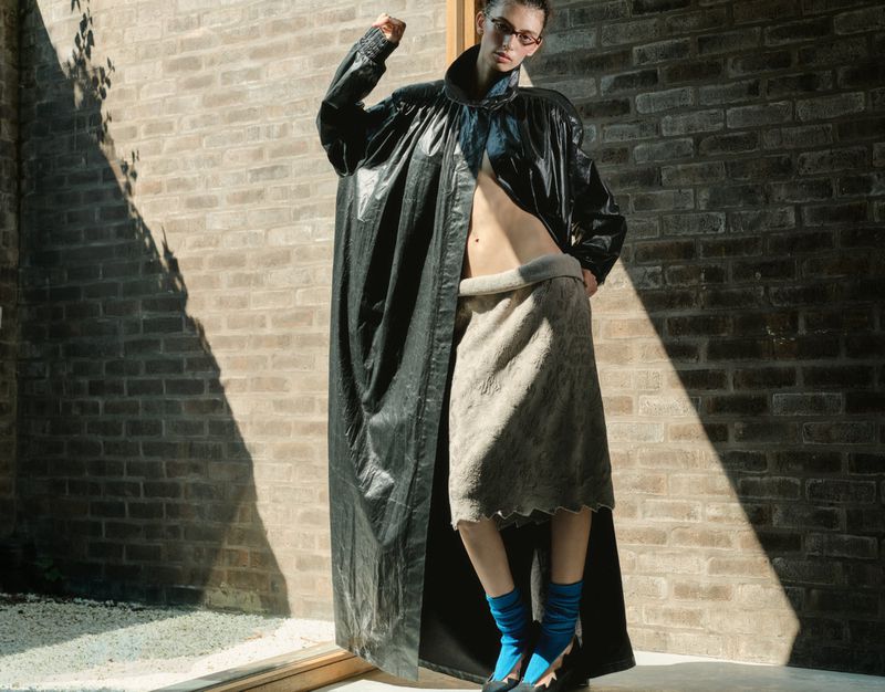 Model wearing an oversized glossy black coat, textured grey skirt, and bright blue socks posing against a sunlit brick wall.