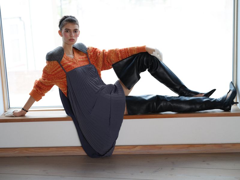 Model reclining along a wooden window ledge wearing an orange patterned sweater, grey pleated pinafore, and black thigh-high leather boots.