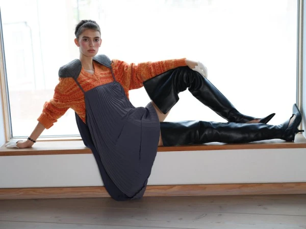 Model reclining along a wooden window ledge wearing an orange patterned sweater, grey pleated pinafore, and black thigh-high leather boots.