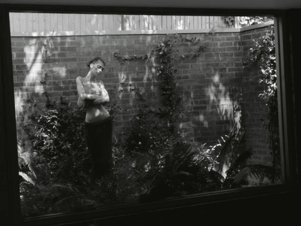 Monochrome portrait of a woman standing in a garden courtyard with brick walls, viewed through a dark window frame.